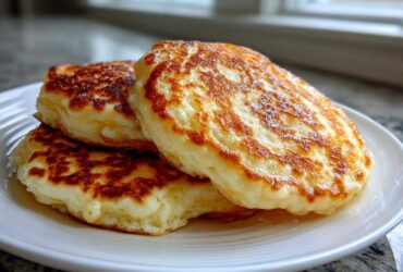 A close-up of three fluffy pancakes stacked on a white plate, showing golden brown, perfectly cooked surfaces.