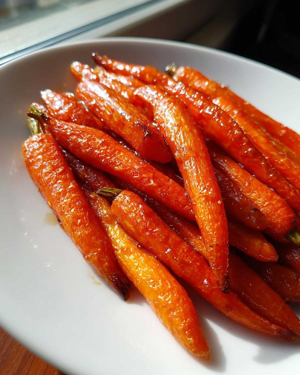 A close-up of bright orange Glazed Honey Garlic Butter Roasted Carrots piled on a white dish, glistening under natural light.