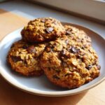 Close-up of four thick, oatmeal-based Go Bananas Cookies studded with dark chocolate chunks on a light plate.