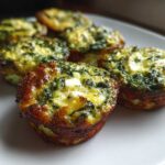 A close-up of several golden brown Breakfast Mini Quiches, featuring visible spinach and white cheese chunks, served on a white plate.