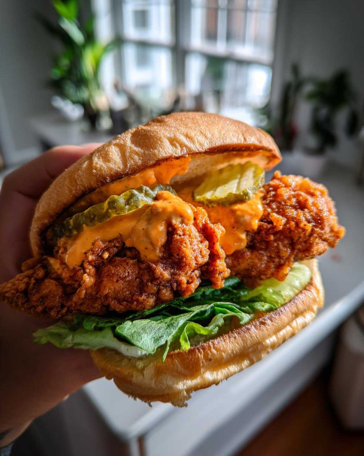 A hand holds a perfectly assembled Crispy Chicken Sandwich featuring a thick piece of fried chicken, spicy sauce, pickles, and lettuce on a brioche bun.