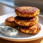 A stack of three golden-brown, crispy Irresistible Salmon Patties served with a dollop of creamy dill sauce on a white plate.
