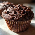 A close-up of a moist, dark chocolate muffin topped generously with chocolate chips, featuring a rich texture.