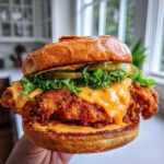 A close-up of a juicy Crispy Chicken Sandwich held by hand, featuring a thick fried patty, orange sauce, pickles, and lettuce on a brioche bun.