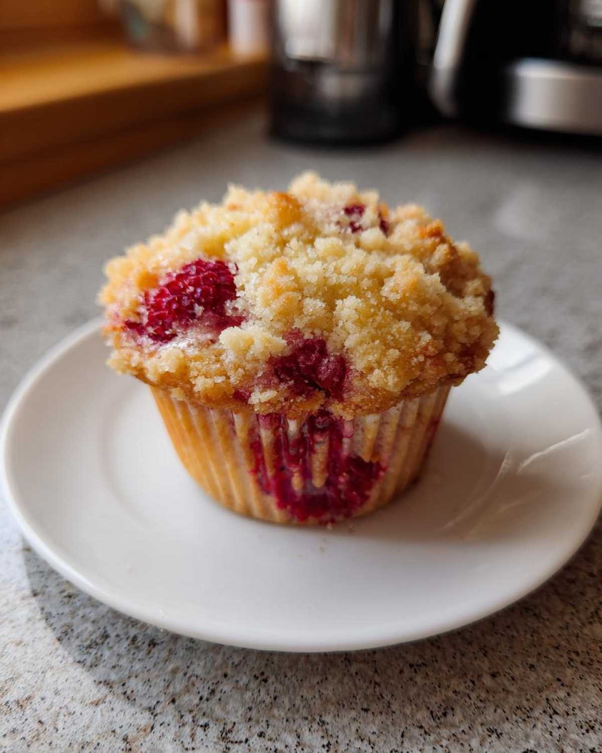 A close-up of a freshly baked Raspberry Streusel Muffins topped with golden, buttery crumbs, sitting on a small white plate.