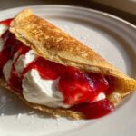 A golden-brown Raspberry Vanilla Crepes folded over a filling of white cream and bright red raspberry sauce.