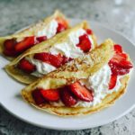 Three folded Strawberry Crepes with Whipped Cream and sliced strawberries, lightly dusted with powdered sugar.