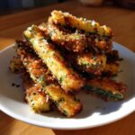 A close-up of crispy, golden Baked Parmesan Zucchini Fries piled high on a white plate, sprinkled with cheese and herbs.