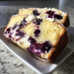 Two close-up slices of moist Blueberry Cream Cheese Bread Loaf showing bright yellow cake, purple blueberries, and white cream cheese swirls.