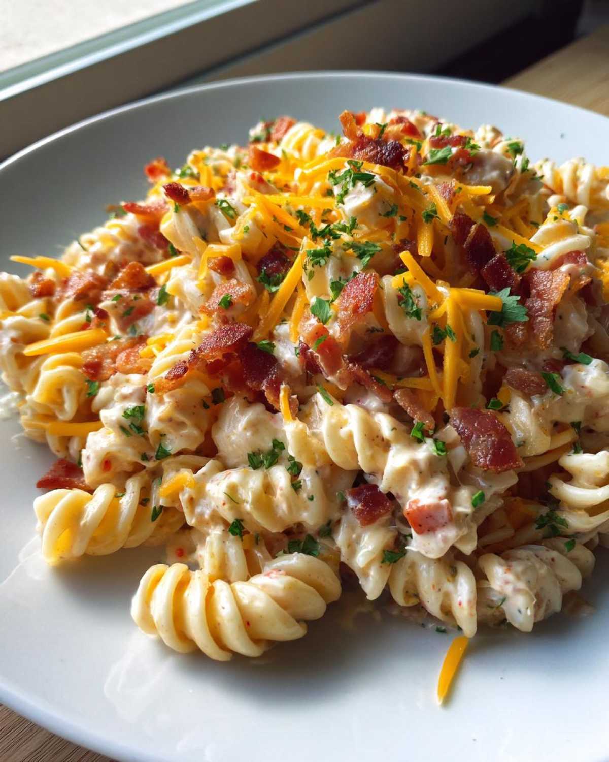 A close-up serving of creamy Bacon Pasta Salad featuring rotini pasta, topped with crumbled bacon, shredded cheddar cheese, and parsley.
