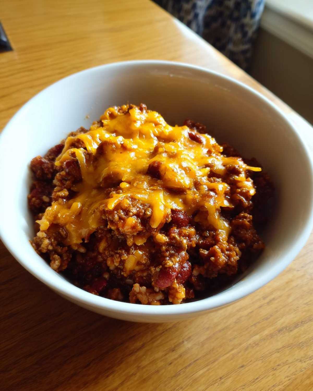 A close-up of a hearty serving of Crock Pot Cowboy Casserole topped with melted cheddar cheese in a white bowl.