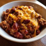 Close-up of a serving of Crock Pot Cowboy Casserole in a white bowl, topped with melted cheddar cheese and visible kidney beans.