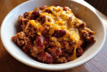 Close-up of a serving of Crock Pot Cowboy Casserole in a white bowl, topped with melted cheddar cheese and visible kidney beans.