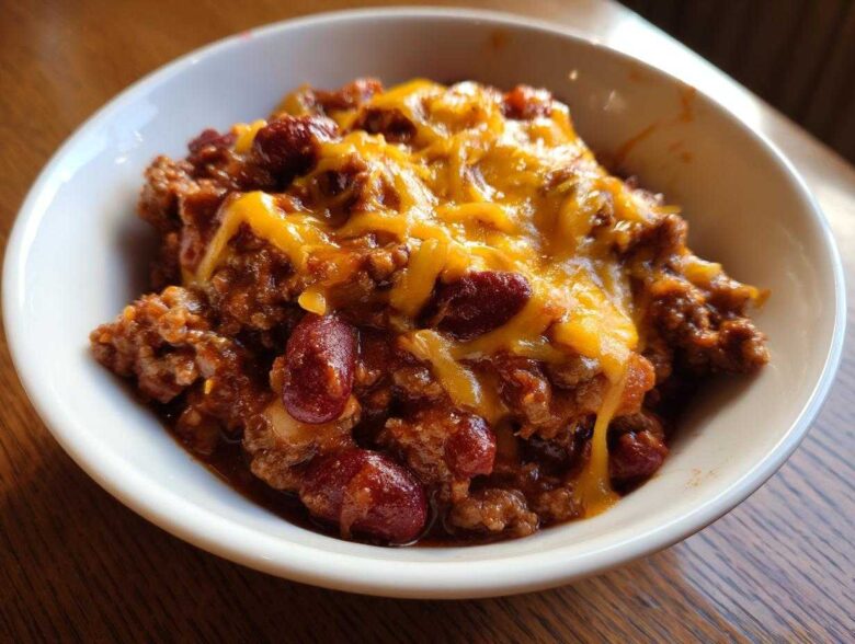 Close-up of a serving of Crock Pot Cowboy Casserole in a white bowl, topped with melted cheddar cheese and visible kidney beans.