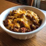 A close-up of a serving of Crock Pot Cowboy Casserole topped generously with melted cheddar cheese in a white bowl.