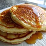 Close-up of a stack of three fluffy Buttermilk Pancakes drizzled generously with golden syrup.