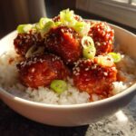 Close-up of glossy, glazed meatballs served over white rice, garnished with sesame seeds and sliced green onions in a white bowl.