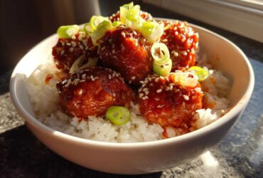 Close-up of glossy, glazed meatballs served over white rice, garnished with sesame seeds and sliced green onions in a white bowl.