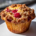 A close-up, brightly lit photo of a single, golden-brown Raspberry Streusel Muffins topped with generous brown sugar streusel and fresh raspberries.