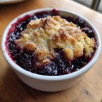 Close-up of a warm, individual serving of Blackberry Cobbler in a white ramekin, featuring bubbling fruit and a golden, sugared topping.